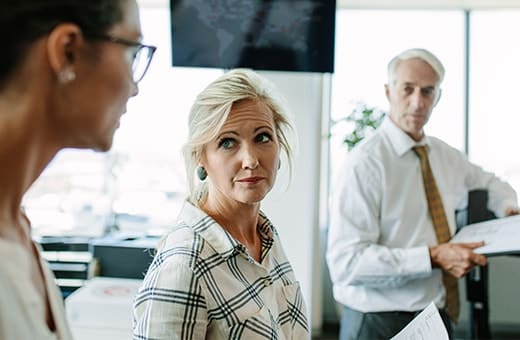 Blond woman in plaid shirt looks toward camera as two coworkers discuss in a bright office, one holding papers.