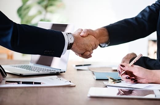 Two professionals shake hands across a conference table, signaling an agreement, with a laptop and documents on the desk nearby.