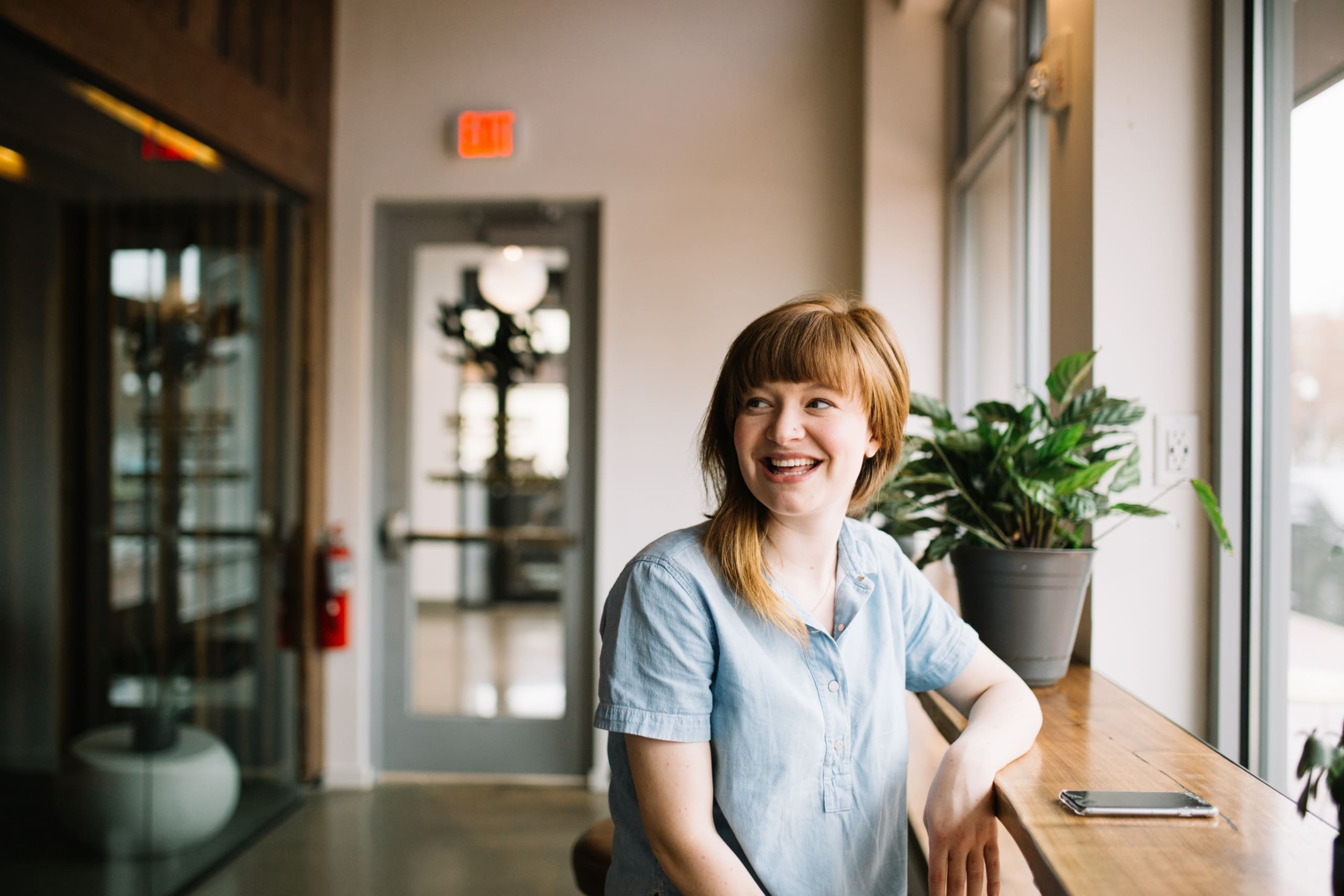 Smiling woman with light blue shirt sitting at a wooden counter by a large window in a bright indoor space.