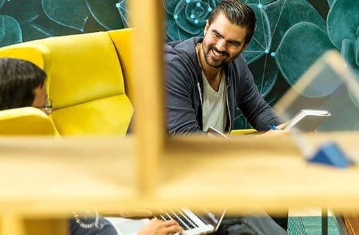 Two people in a bright lounge with yellow chairs, one using a laptop and the other holding a tablet.