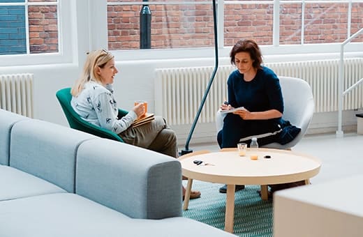 Two women sit across from each other in a bright, modern room; a therapist takes notes while the client speaks, seated on comfortable chairs with a round table between them.