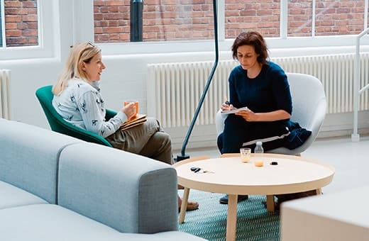 Two women in a bright room having a counseling session: one holds a mug, the other writes notes at a round coffee table.
