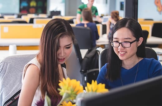 Two young women collaborate at a computer in a bright open workspace, with yellow partitions and flowers on the desk in the foreground.