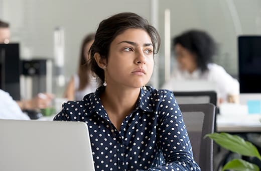 Woman in a navy polka-dot shirt sits at a desk in a bright office, looking off to the side with a thoughtful expression.