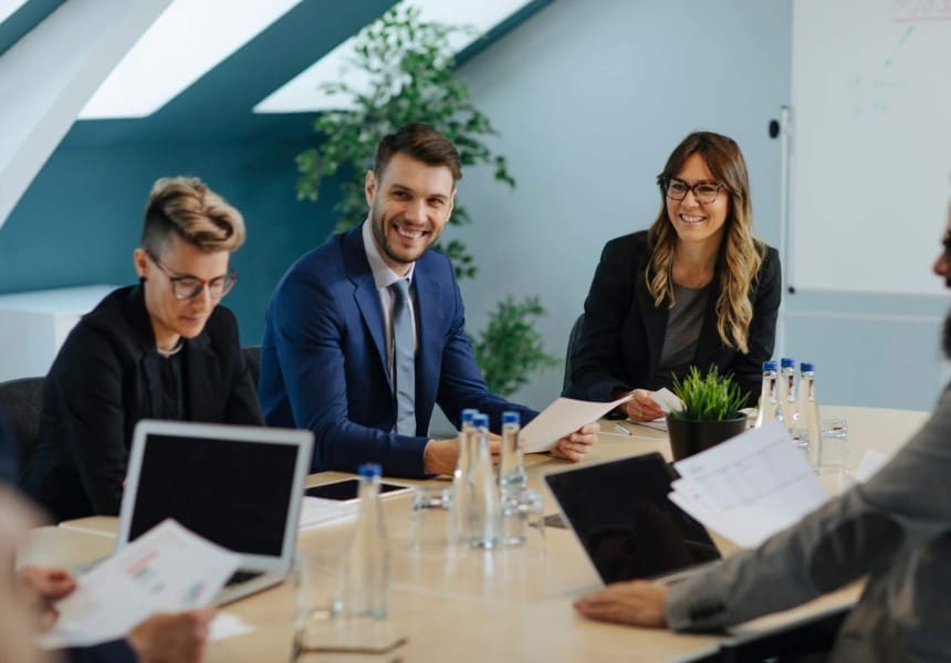 Team of professionals in a business meeting around a table with laptops and papers, smiling and collaborating.