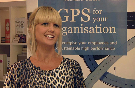 Smiling woman with blonde hair wearing a leopard-print top, standing in front of a blue banner that says 'GPS for your organisation'.