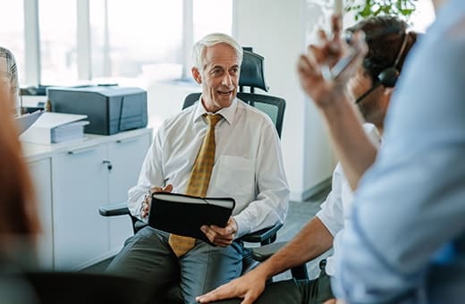 Older man in a white shirt and yellow tie holds a tablet, engaging in a business meeting with colleagues wearing headsets in a bright office.