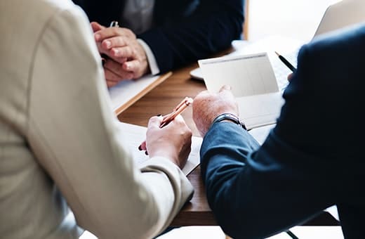 Two professionals signing documents at a wooden table during a meeting with notes and pens nearby.