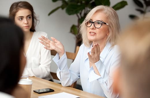 Older woman with glasses speaks and gestures at a meeting table, as a younger woman listens nearby.