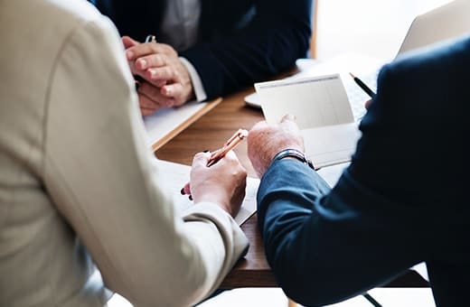 People in business attire sign a document on a wooden table, with multiple hands and pens visible during a meeting.