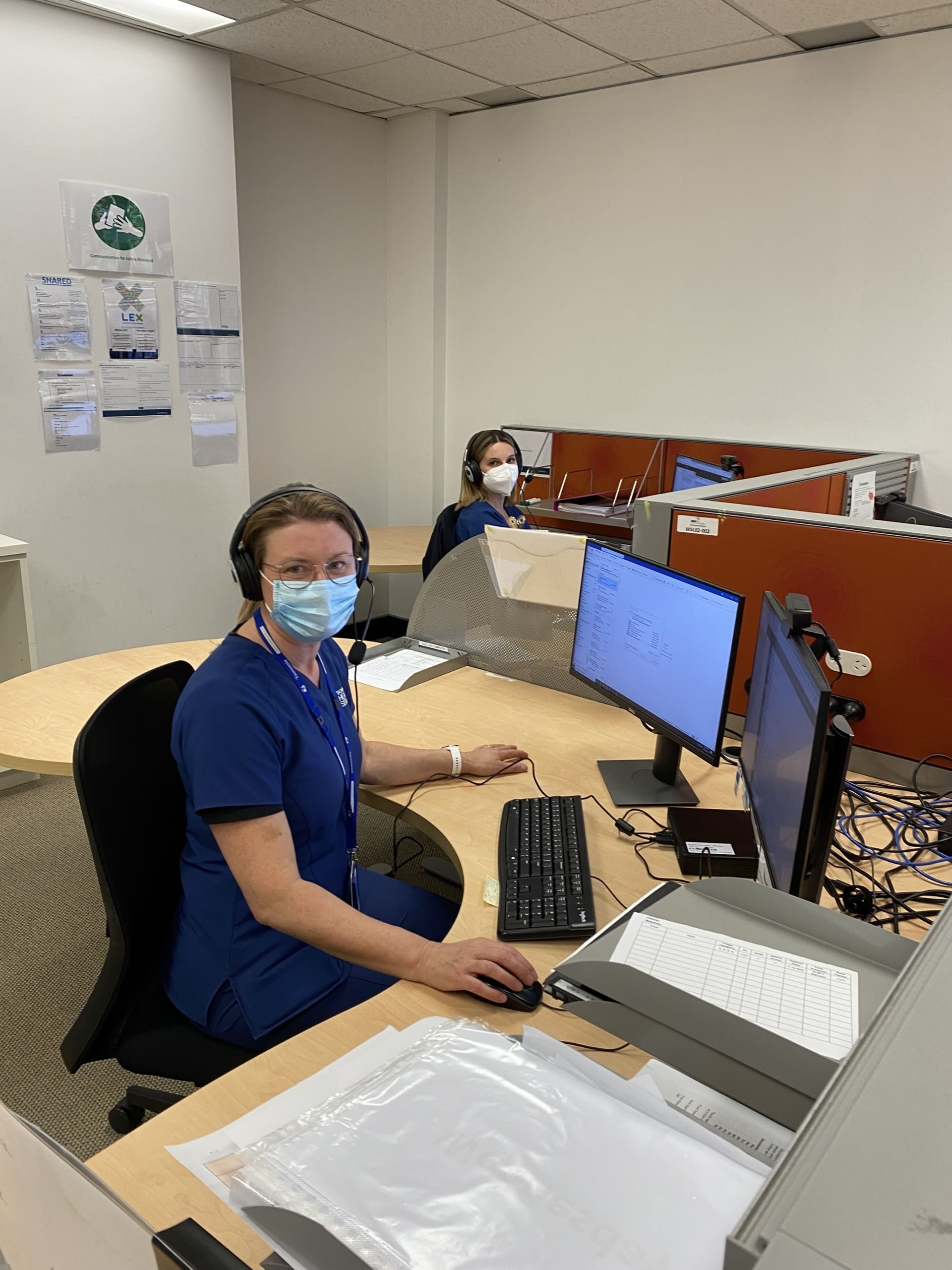 Two healthcare workers in blue scrubs wearing masks and headsets sit at computer desks in an office workspace with cubicle dividers
