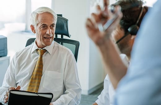 Senior man with white hair in a white shirt and yellow tie speaks with a colleague wearing a headset in a modern office
