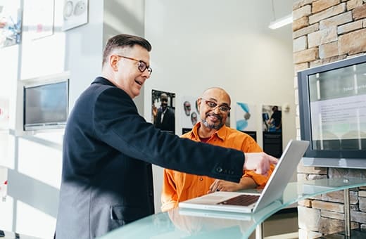 Two men in a showroom examining a laptop on a glass counter; one in a dark suit points at the screen while the other in an orange shirt smiles.