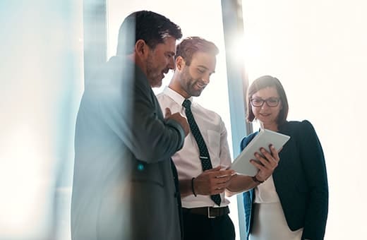 Three professionals in business attire gather around a tablet, discussing work in a bright office hallway.
