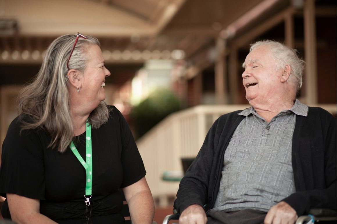 Woman with gray hair and a green lanyard sits and smiles at an elderly man in a wheelchair as they chat outdoors under a covered area.