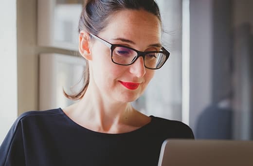 Woman with dark hair and glasses working on a laptop in a bright office environment with a focused expression on her face in natural light from a window.