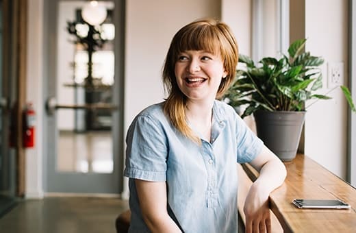 Woman with light brown hair smiling, sitting at a window counter in a bright office, wearing a light blue shirt with a tablet nearby.