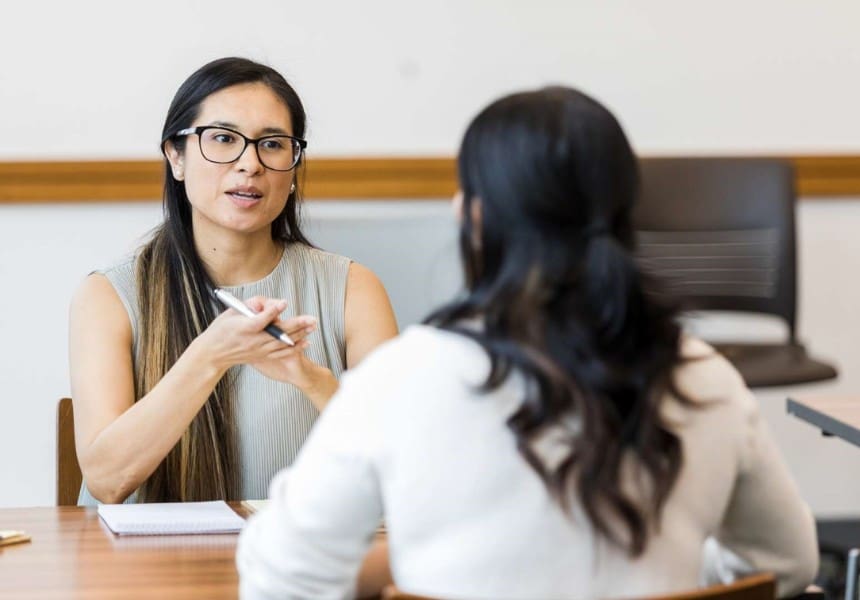 Two women sit across from each other at a desk in an office; one wears glasses and speaks with a pen in hand.