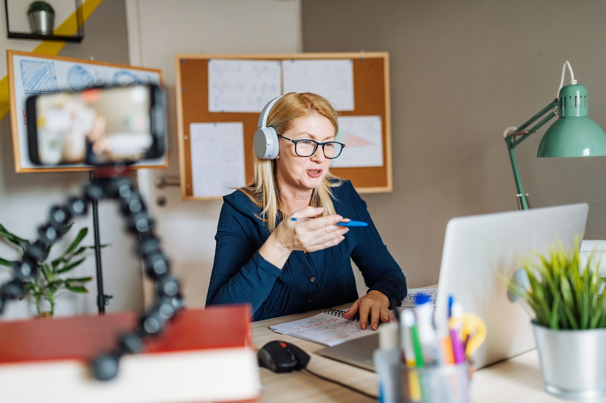 Blonde woman wearing headphones and glasses speaks while gesturing at a desk cluttered with papers and a laptop in a home office set-up.