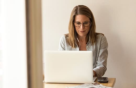 Woman wearing glasses works on a white laptop at a wooden desk, with papers and a phone nearby.