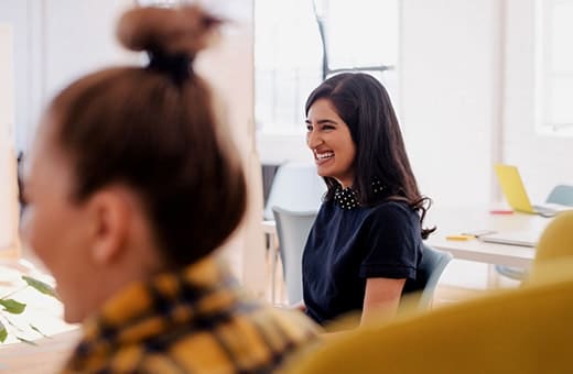 Smiling woman with dark hair in a navy top sits in a bright office, chatting with a colleague.