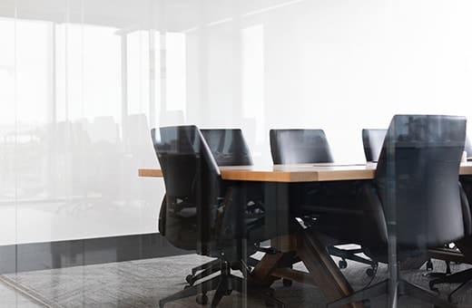 Modern conference room with a wooden table and black swivel chairs around it, surrounded by glass walls and natural light.