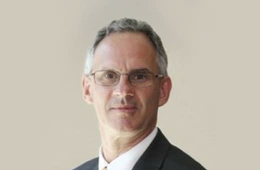 Professional headshot of a middle-aged man in a dark suit and glasses against a neutral background.