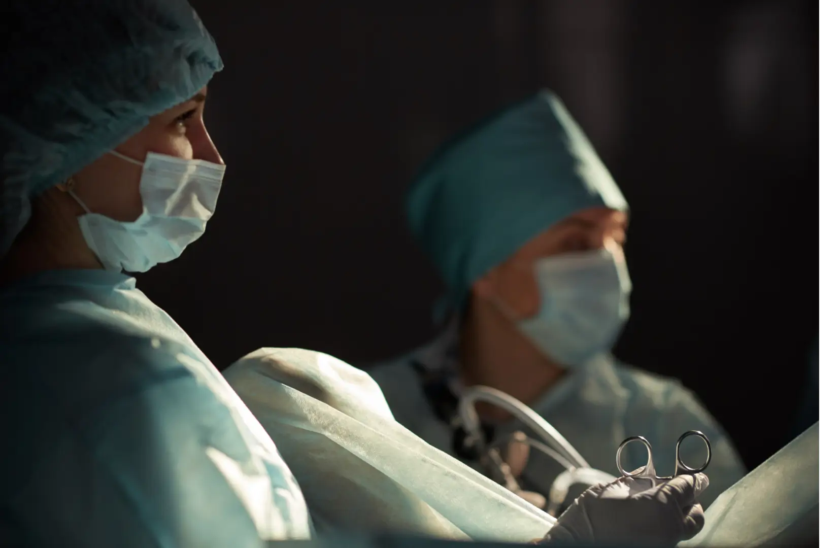 Two surgeons in blue scrubs, masks, and caps perform surgery in an operating room, one holding surgical forceps.