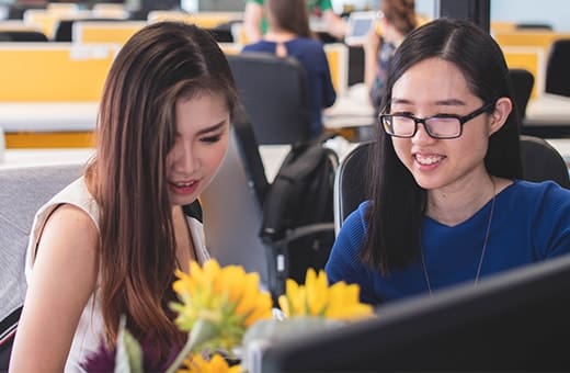 Two young women sit together at a computer, smiling as they look at the screen, with yellow chairs and flowers on the desk nearby.