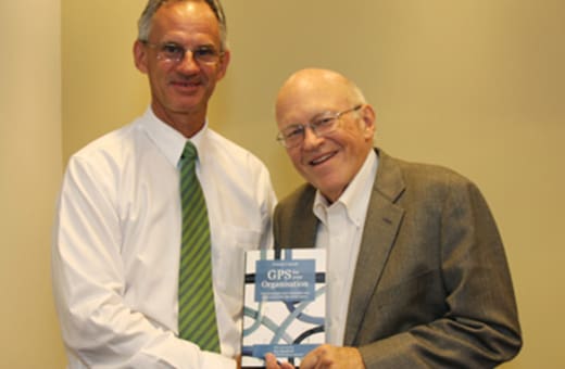 Two men in business attire shake hands indoors while holding a booklet facing the camera.