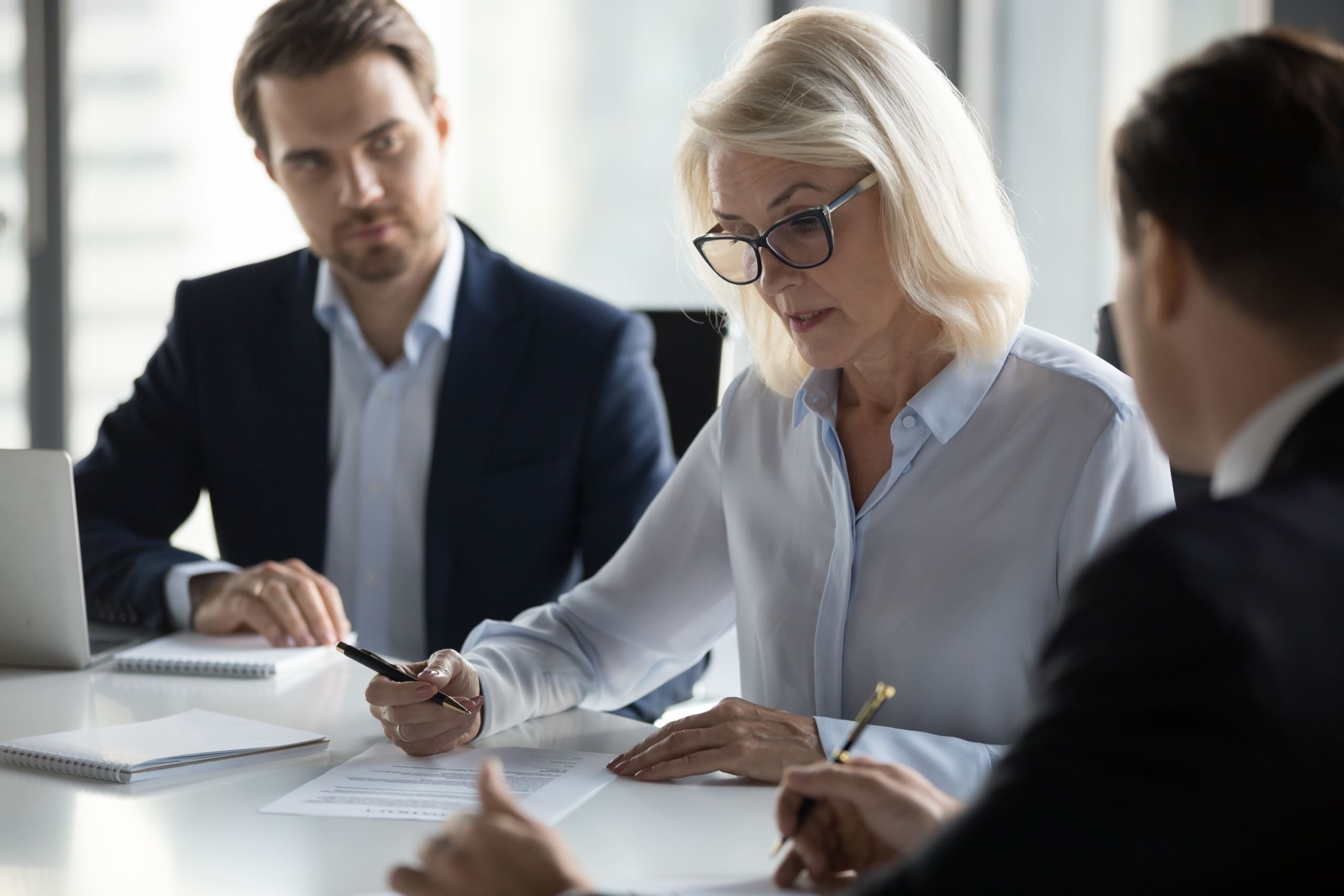 Business professionals in a meeting around a white table, reviewing documents with pens and a laptop visible in the background.