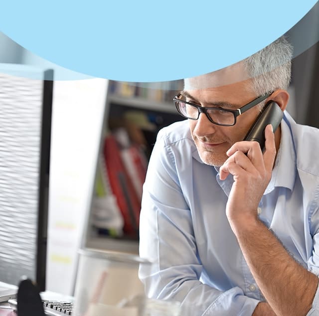 Middle-aged man with gray hair and glasses talking on a mobile phone at a desk in a modern office, computer monitor visible in the background.