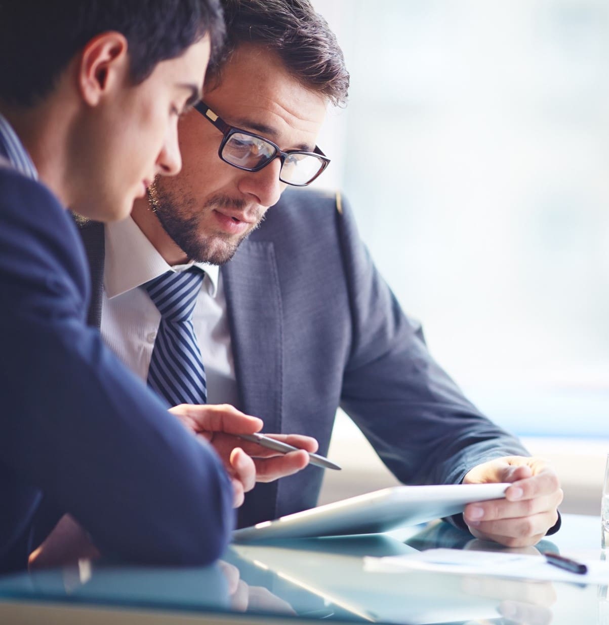 Two businessmen in suits collaborate over a tablet at a glass desk, one holding a pen.