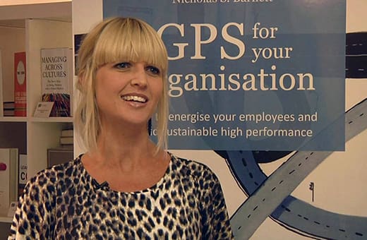 Professional woman with blonde hair in a leopard-print top speaking at a presentation; behind her is a blue banner reading 'GPS for your organisation' and 'Energise your employees and sustainable high performance' (text partly visible).