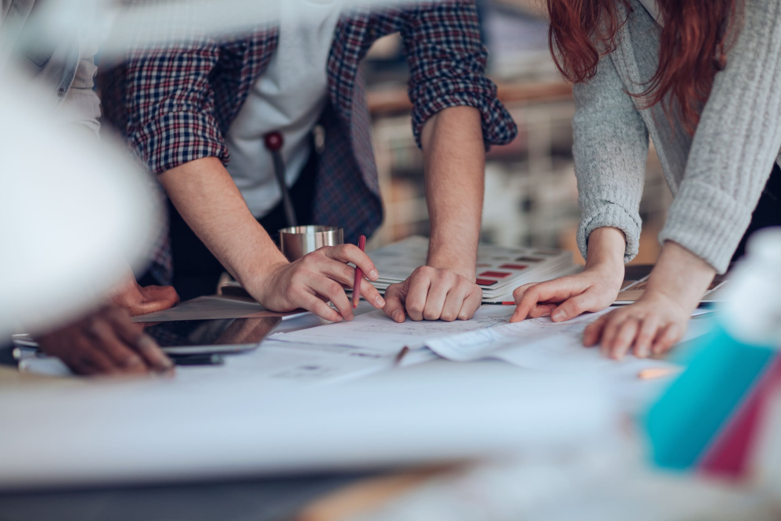Group of people leaning over a table, drawing and annotating large sheets of paper with pencils and markers in a collaborative planning session.