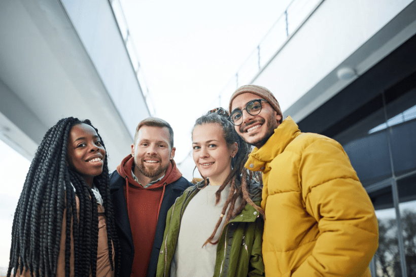 Four diverse friends posing together outdoors, smiling near a modern architectural backdrop.