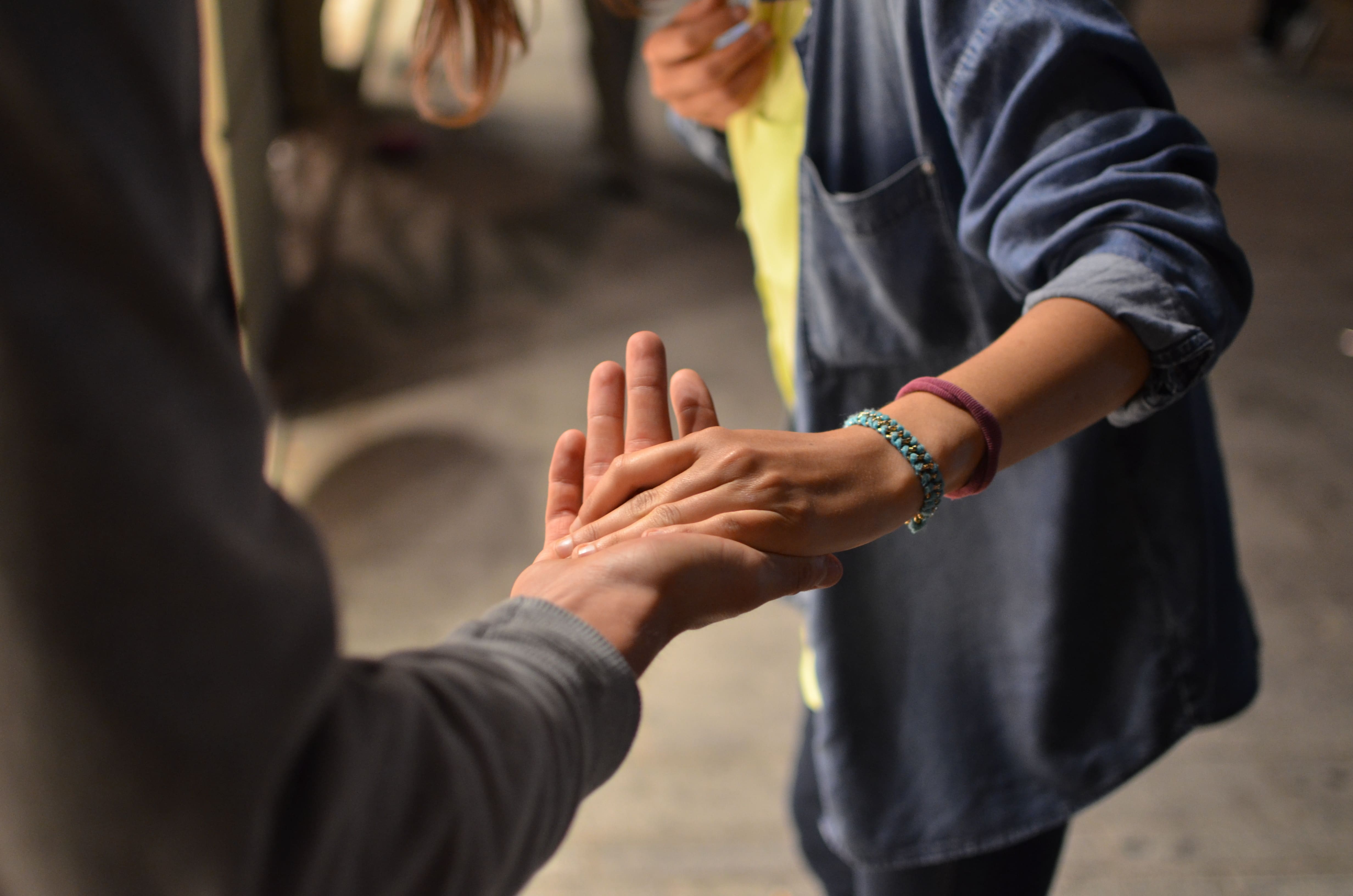 Two people grasp each other’s hands in a friendly handshake/high-five, one wearing a blue denim shirt and bracelets around the wrist.