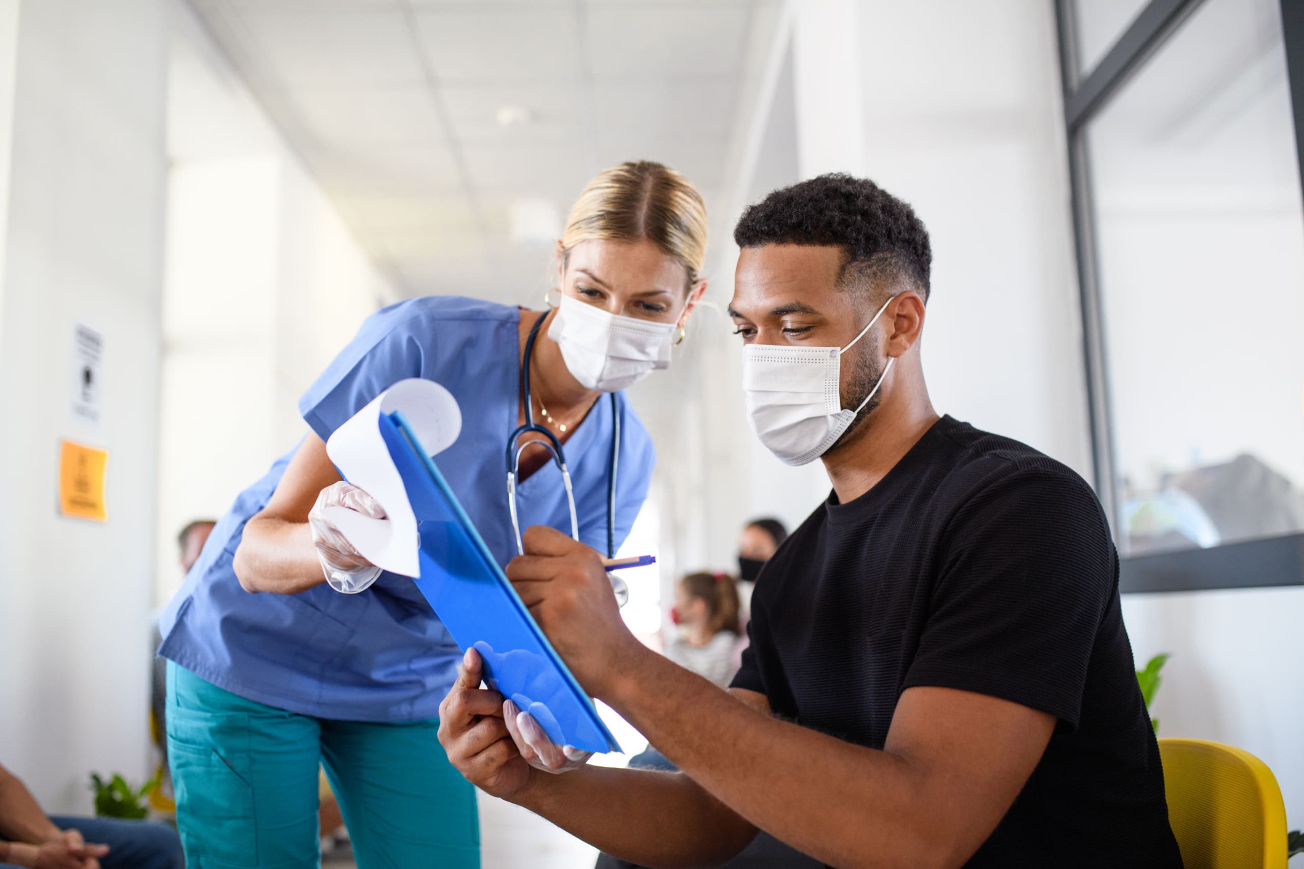 Healthcare worker in blue scrubs and mask assists a man with signing a clipboard in a clinic hallway