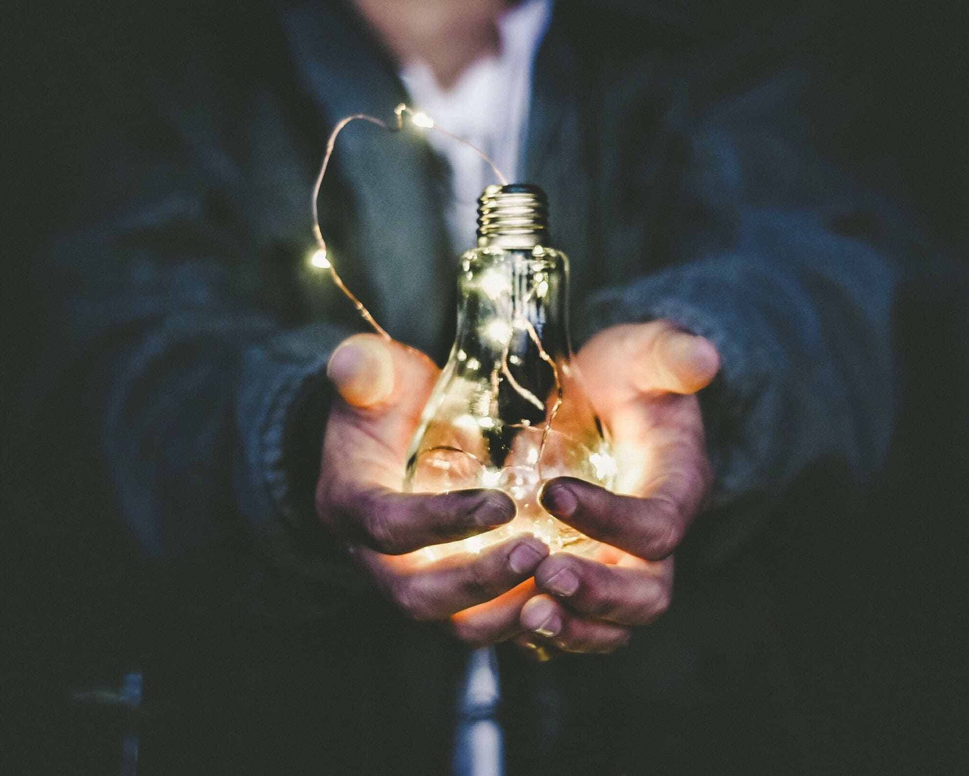 Hands cradle a glowing light bulb wrapped with string lights in a dark setting.