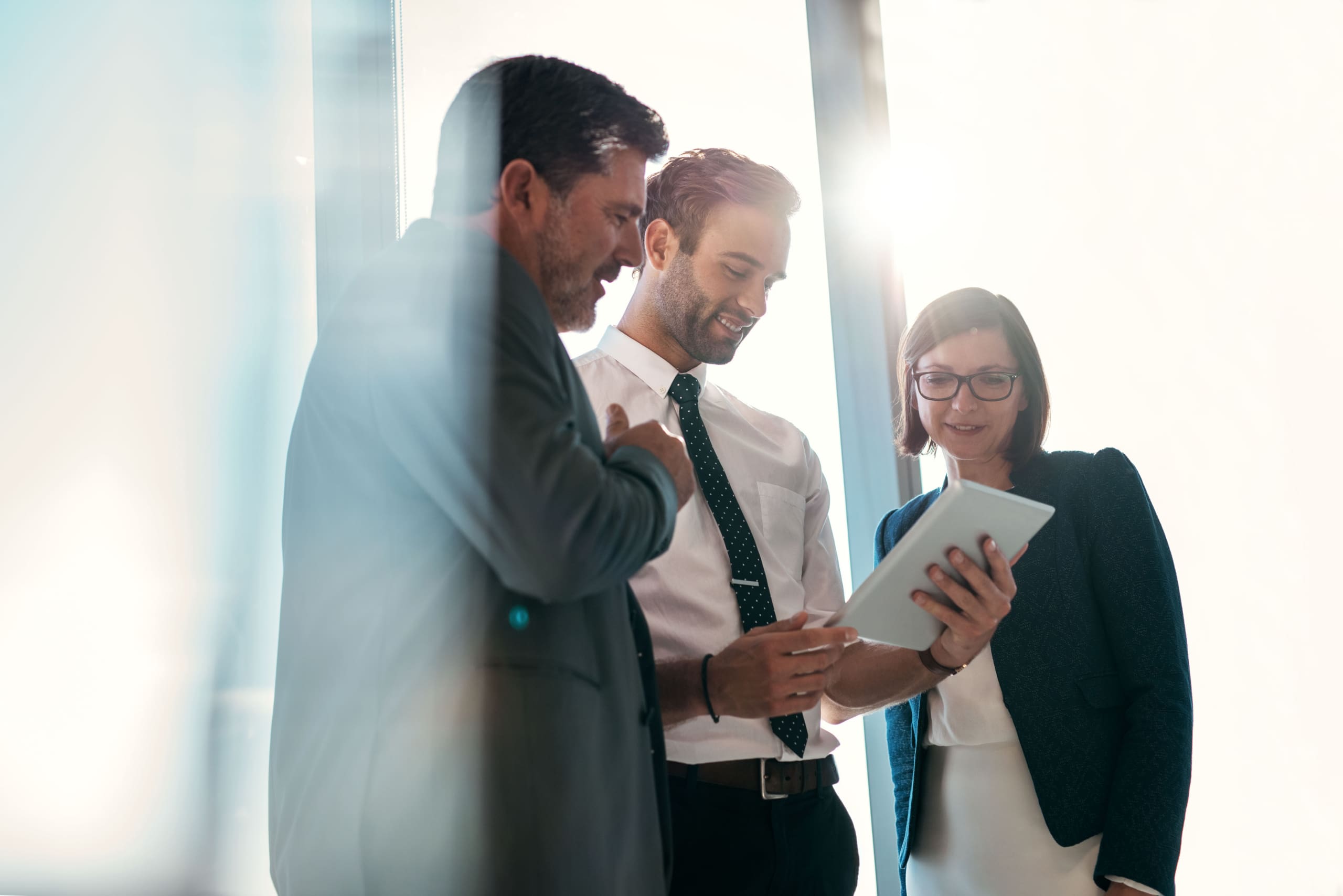 Three professionals stand together in a bright office, looking at a tablet and discussing something written on the screen/board side by side near a glass wall.