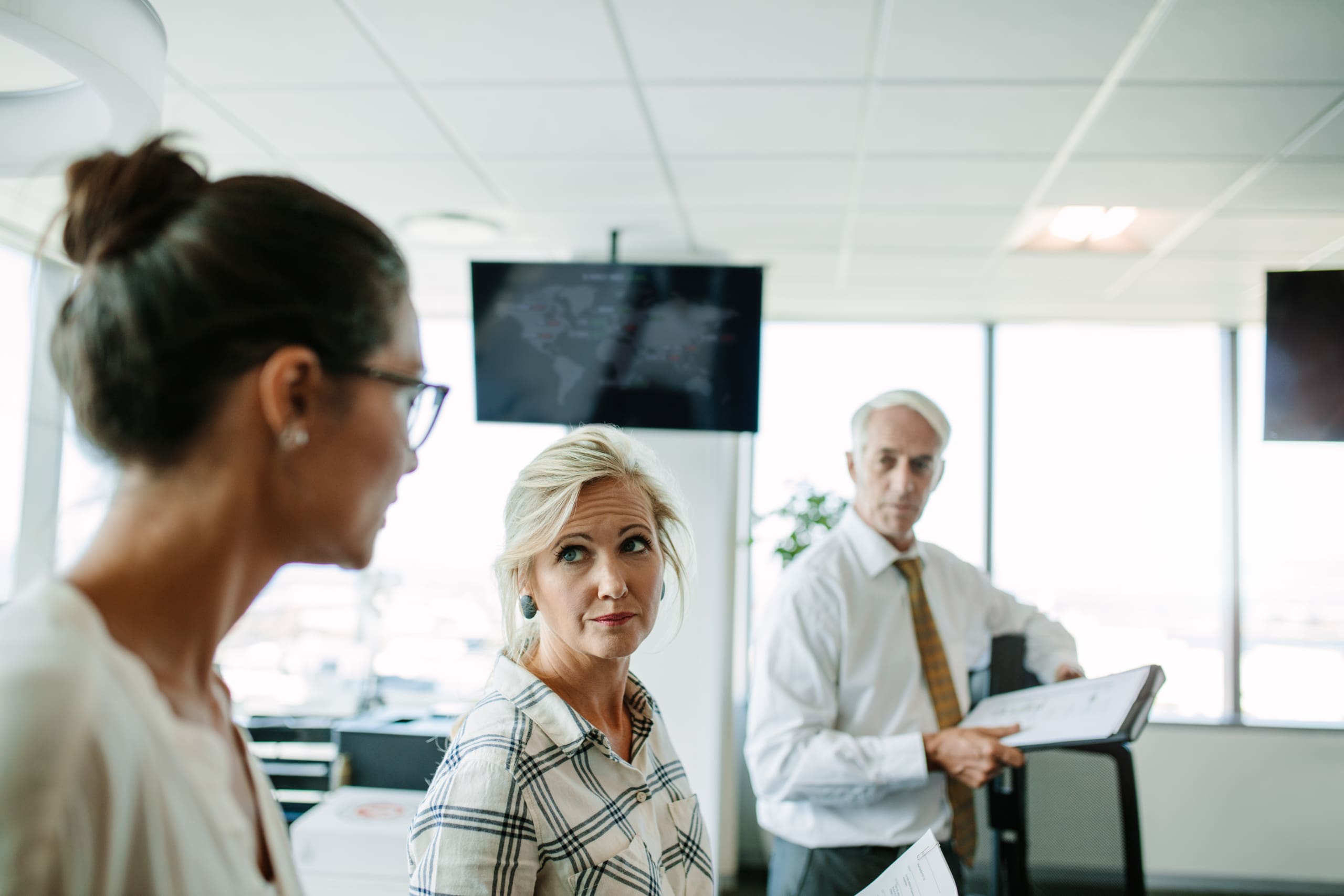 Blonde woman with a headset in the foreground, looking at the camera; two coworkers in a bright office behind her.