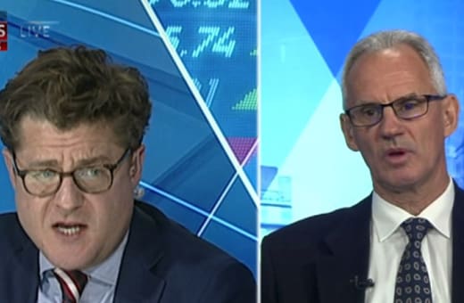 Split-screen of two male news presenters in suits; left presenter with glasses speaks, right presenter with gray hair and glasses looks at the camera, blue geometric studio background.
