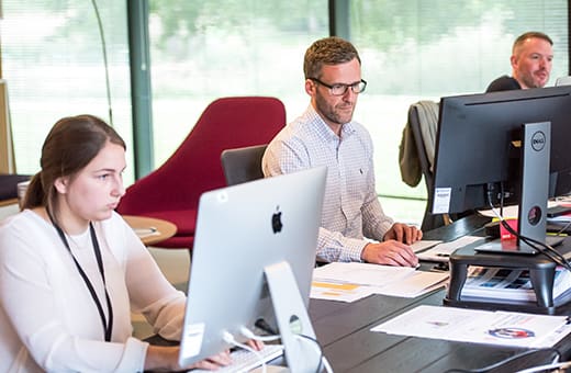 Office scene with three coworkers at desks using desktop computers and documents on the table.