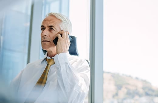 Older man in a white shirt and yellow tie talks on a cellphone by a window in an office setting.